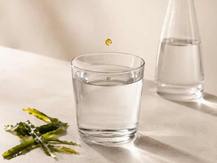 Ein goldener Tropfen fällt in ein Glas Wasser, daneben liegen Algenblätter